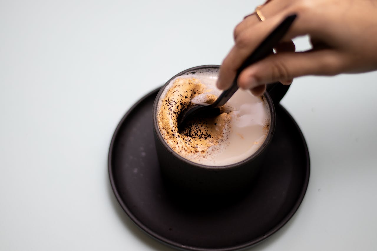 A hand stirring a foamy coffee in a black cup on a saucer.
